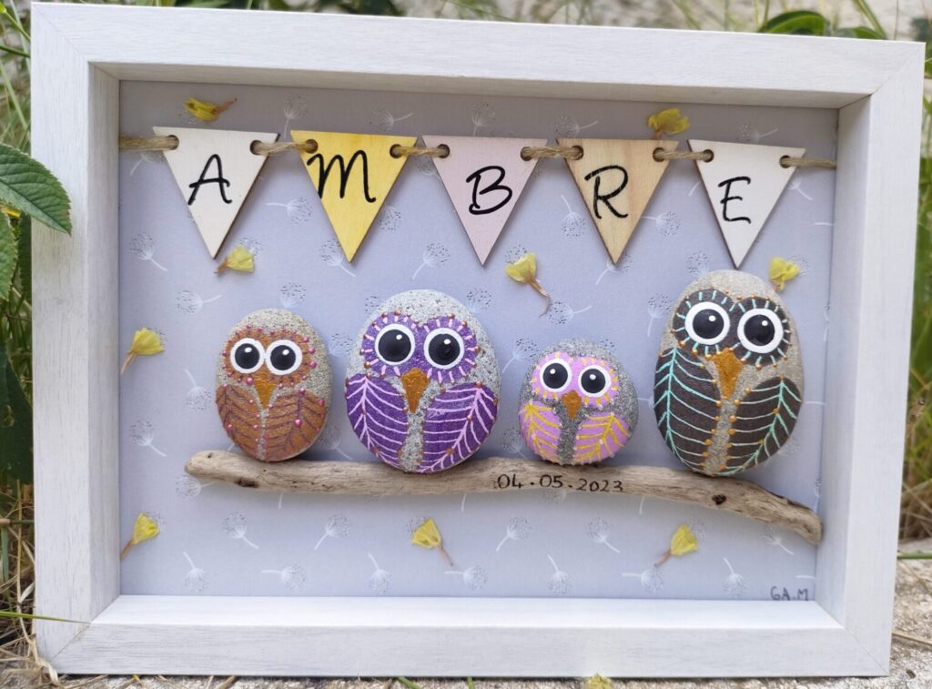 cadre de naissance 4 galets peints en forme de chouettes (papa, maman et deux enfants) sur branche de bois flotté avec date de naissance du dernier bébé né inscrite dessus). Petite guirlande de fanions au dessus du galets au prénom de l'enfant ici AMBRE. cadre format moyen couleur bois naturel et fond gris à motif fleural